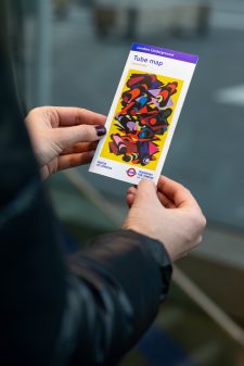 The 33rd edition of the pocket tube map cover by Phyllida Barlow, a bright yellow artwork overlaid with colourful geometric shapes mimicking tube tunnels, in the hand of a member of the public.