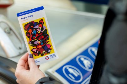 The 33rd edition of the pocket tube map cover by Phyllida Barlow, a bright yellow artwork overlaid with colourful geometric shapes mimicking tube tunnels, in the hand of a member of the public.