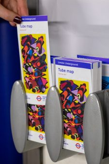 A hand picks a copy of the 33rd edition of the pocket Tube map cover by Phyllida Barlow, a bright yellow artwork overlaid with colourful geometric shapes mimicking tube tunnels, from racks in a station.