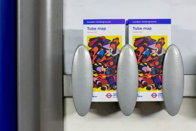 The 33rd edition of the pocket tube map cover by Phyllida Barlow, a bright yellow artwork overlaid with colourful geometric shapes mimicking tube tunnels, in racks in a station.