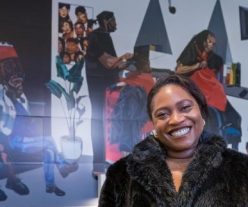 A portrait of British-Nigerian artist Joy Labinjo smiling in front of her artwork at Brixton, a scene in a black hairdresser, a mass of blacks, greys, greens and orange behind her.