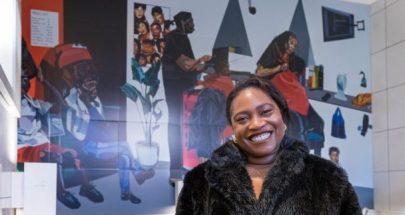 A portrait of British-Nigerian artist Joy Labinjo smiling in front of her artwork at Brixton, a scene in a black hairdresser, a mass of blacks, greys, greens and orange behind her.